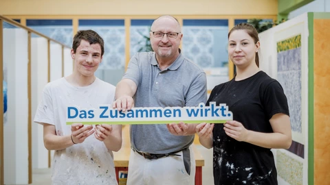 Zwei Auszubildende und ein Ausbilder halten einen Schriftzug "Das Zusammen wirkt".