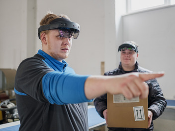 Ein Auszubildender mit einer Mixed-Realitiy-Brille zeigt mit dem Finger auf etwas. Im Hintergrund steht ein anderer Azubi, er trägt auch eine MR-Brille und hält eine Kiste in den Händen.