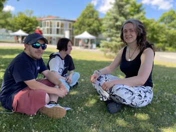 Drei Personen sitzen auf einer Wiese im Freien und unterhalten sich. Auf der rechten Seite sitzt Luisa.