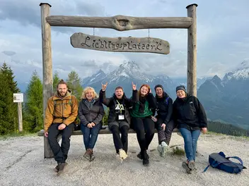 Sechs Personen auf einer Bank mit einem Schild "Lieblingsplatzerl". Im Hintergrund sind Berggipfel.