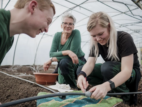 Zwei Auszubildende und eine Ausbildern knein in einem Gewächshaus auf der Erde und arbeiten an einem Bewässerungssystem. 