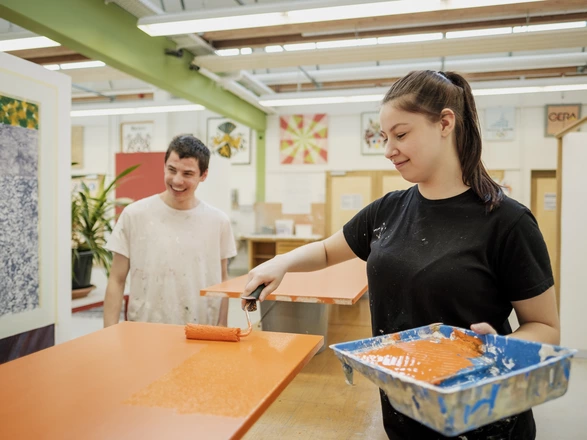 Zwei Auszubildende im Bereich Farbtechnik und Raumgestaltung. Eine junge Frau trägt mit einer Farbrolle orange Farbe auf eine Holzplatte auf.