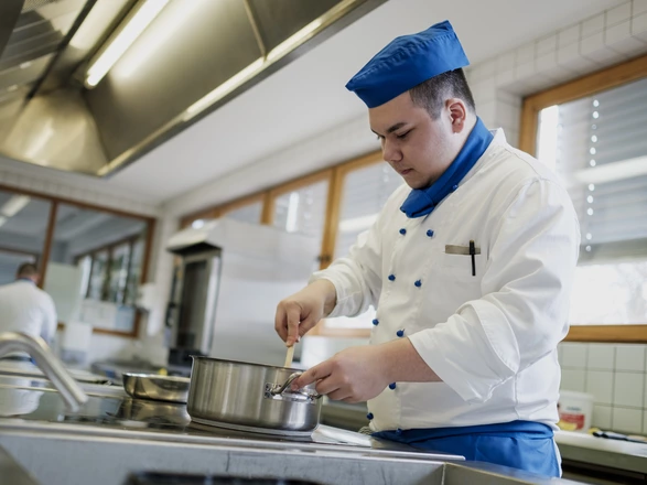 Ein Jugendlicher in Arbeitskleidung rührt in der Ausbildungsküche in einem Topf.