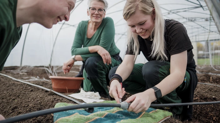 Zwei Auszubildende und eine Ausbildern knein in einem Gewächshaus auf der Erde und arbeiten an einem Bewässerungssystem. 