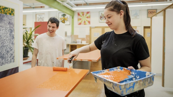 Zwei Auszubildende im Bereich Farbtechnik und Raumgestaltung. Eine junge Frau trägt mit einer Farbrolle orange Farbe auf eine Holzplatte auf.