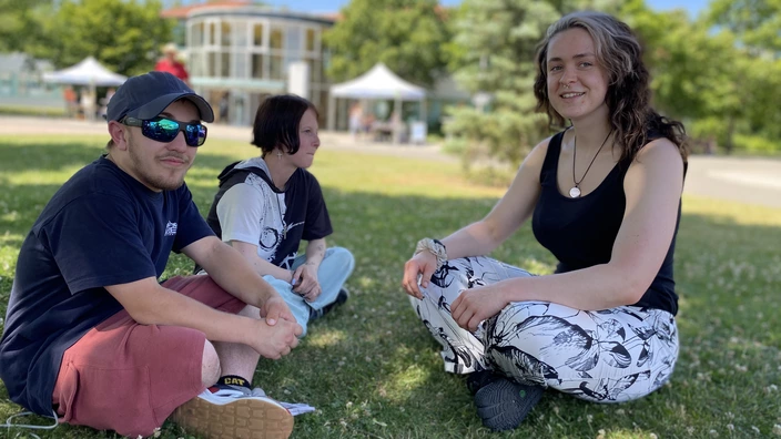 Drei Personen sitzen auf einer Wiese im Freien und unterhalten sich. Auf der rechten Seite sitzt Luisa.