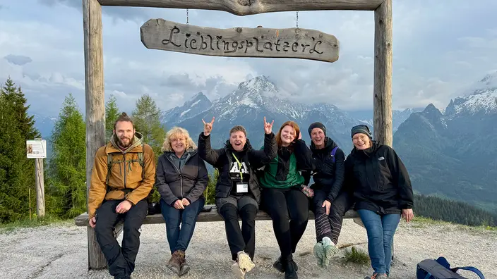 Sechs Personen auf einer Bank mit einem Schild "Lieblingsplatzerl". Im Hintergrund sind Berggipfel.