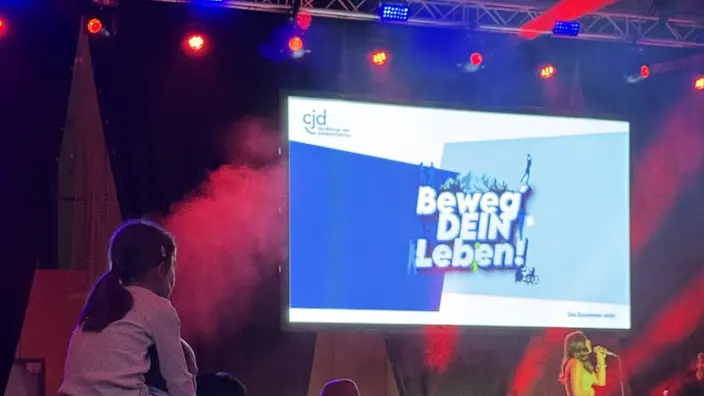 Publikum bei der Eröffnungsveranstaltung. Auf dem Bildschirm über der Bühne steht "Beweg DEIN Leben!"